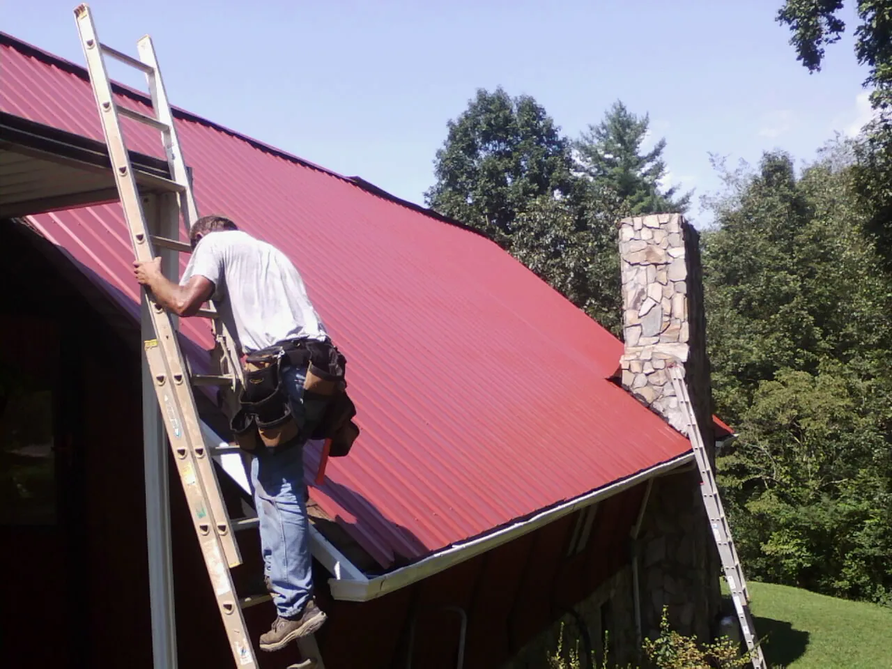 Roofer on a ladder beside a red metal roof and stone chimney — Soffit & Fascia Repair by Roofing Brothers in Obernburg